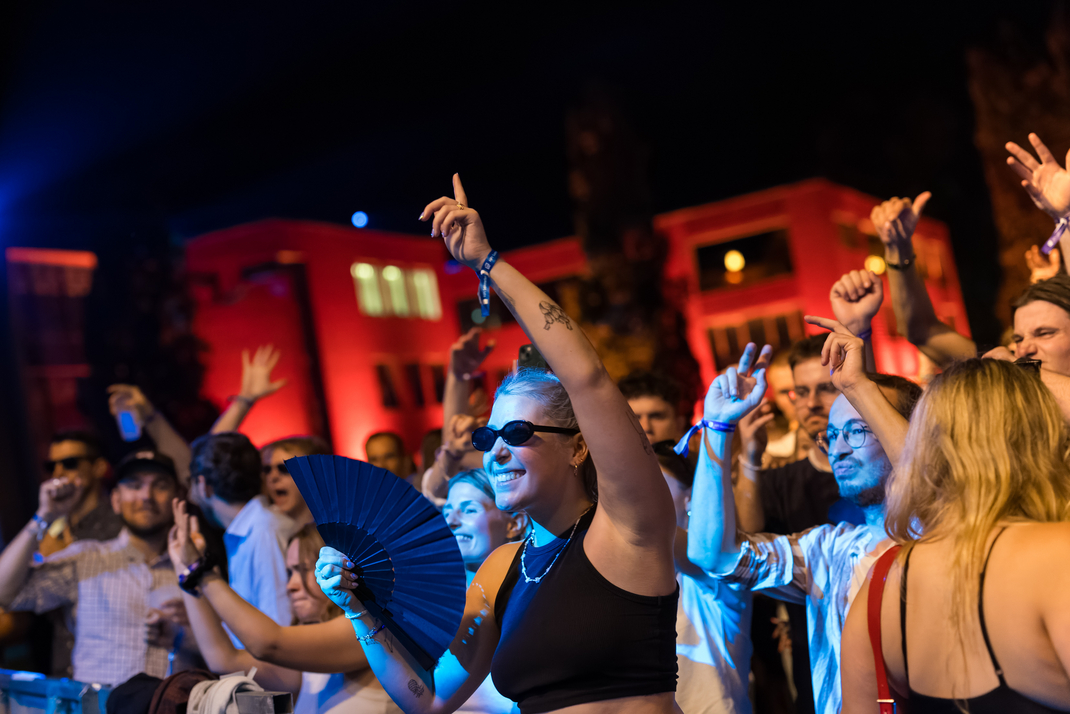  Eine große, ausgelassene Menschenmenge auf dem Sommerfest. Im Vordergrund eine junge Frau mit Sonnenbrille und Fächer, tanzend und lächelnd. Im Hintergrund das HPI-Gebäude, in farbiges Licht getaucht (blau und rot), Nachtstimmung.