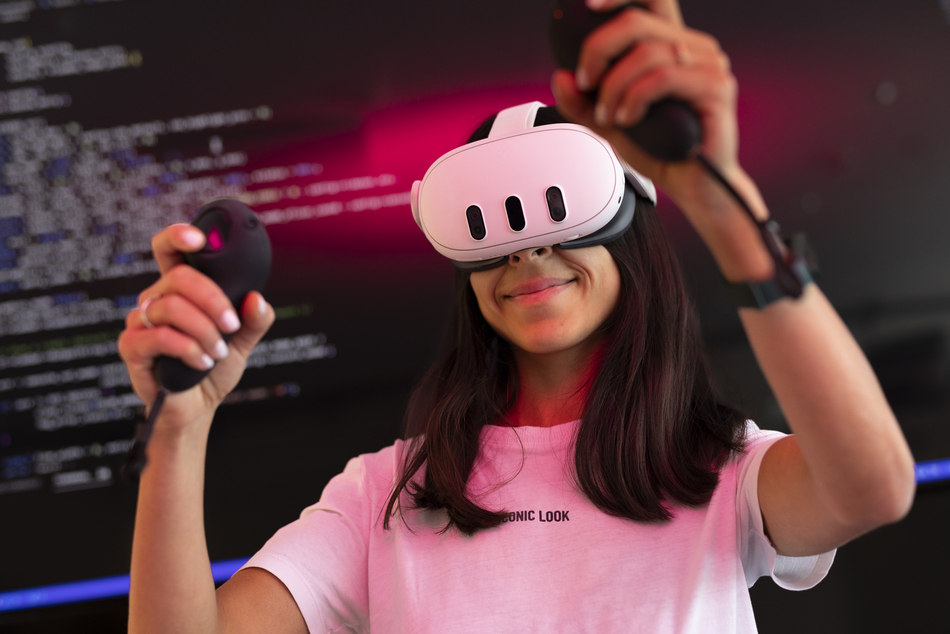 Eine studierende Person trägt eine VR-Brille und hält VR-Controller in den Händen. Im Hintergrund ist ein Display mit Programmcode zu sehen.