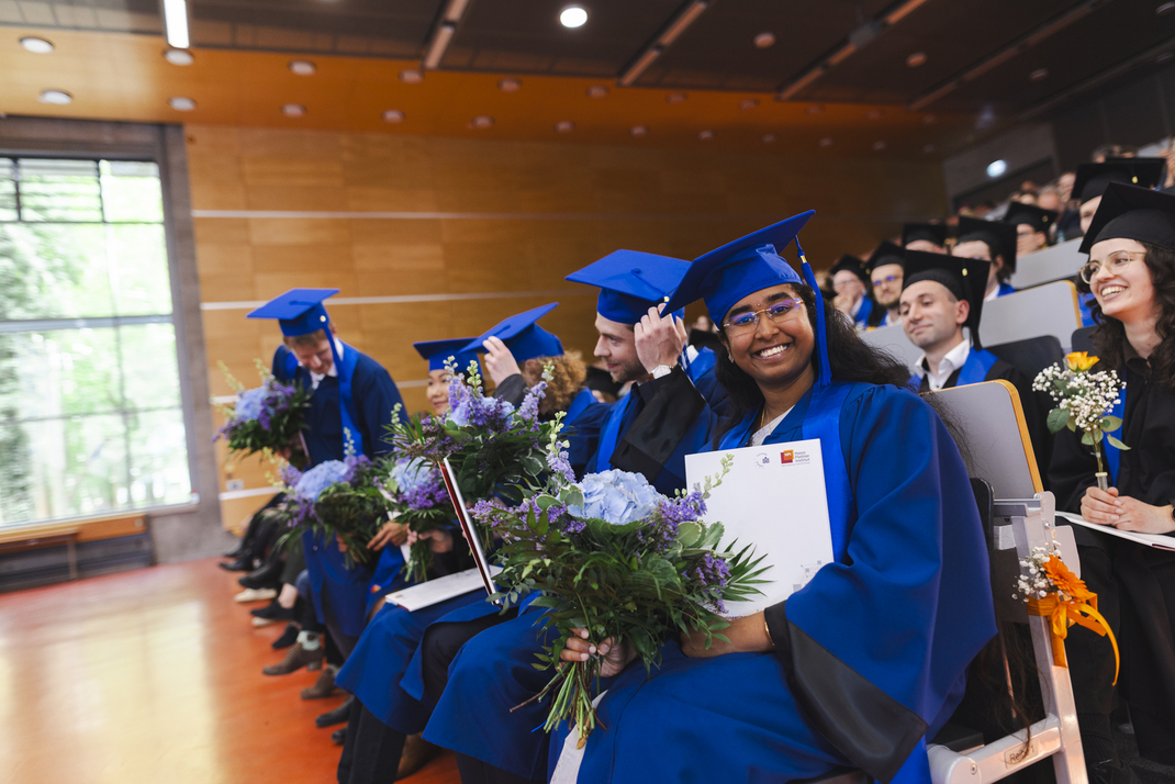 The photo shows proud graduates in the audience, holding their diplomas and bouquets of flowers