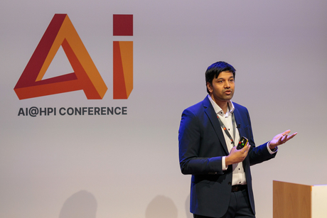 HPi Professor Dr. Gerard de Melo gives a lecture. Behind him is the logo of the AI@HPI conference.