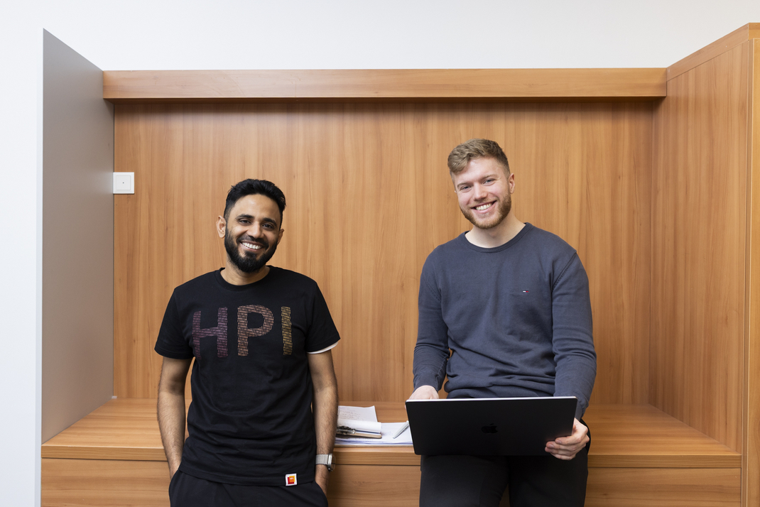 The two HPI Master's students SaadBin Khalid (left) and Pierre Burghardt smile friendly into the camera. SaadBin is wearing a T-shirt with HPI lettering. Pierre holds a laptop in his hands.