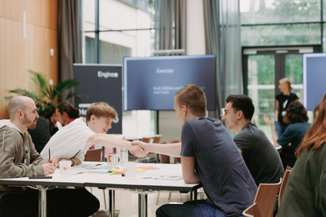 Four participants in the event are working together at a workspace. Two of them are shaking hands.