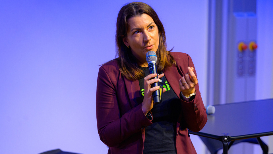 A woman speaks on stage at the Tech Leadership Conference, holding a microphone in one hand and a presentation remote in the other. She wears a burgundy blazer over a black t-shirt and gestures as she addresses the audience.