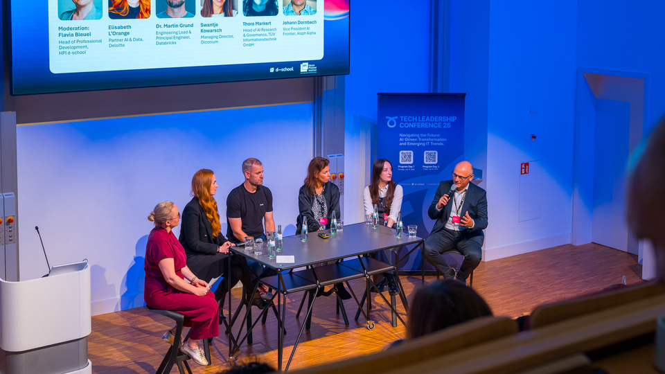 A six-person panel discusses on stage at the Tech Leadership Conference 2025, moderated by Flavia Bleuel (HPI D-School). A projection screen in the background displays the names and roles of the participants, including representatives from Deloitte, Databricks, Diconium, TUI, and Aleph Alpha. A conference banner stands to the right of the stage.