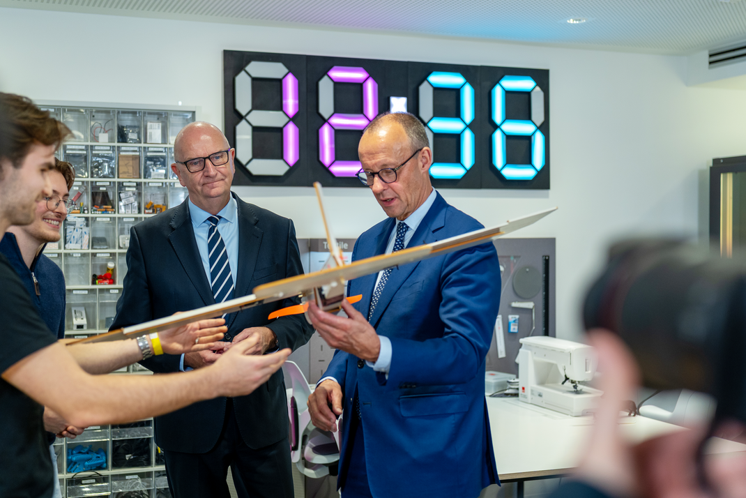 Bundeskanzler Friedrich Merz betrachtet ein Modellflugzeug, das ihm zwei Studierende überreichen. Neben ihm steht Brandenburgs Ministerpräsient Dietmar Woidke.