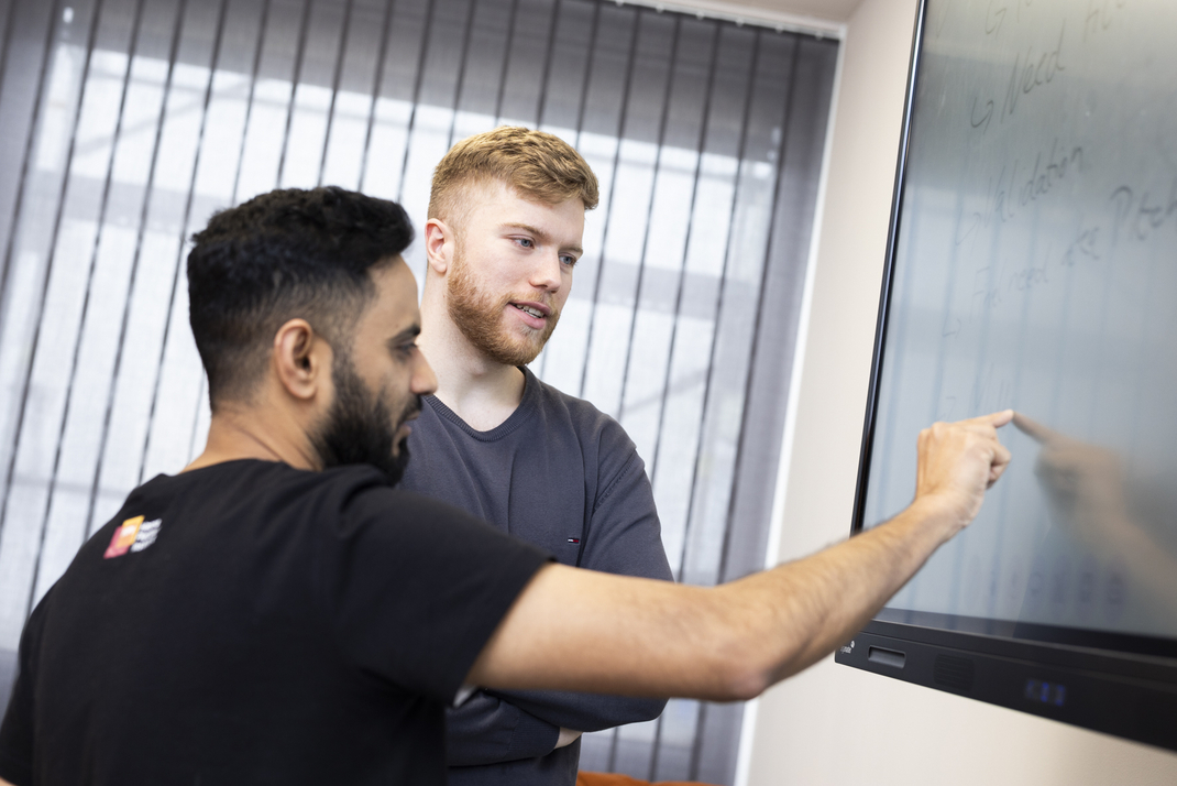 The two HPI master's students SaadBin and Pierre are standing in front of a screen. SaadBin taps on the screen with his index finger.