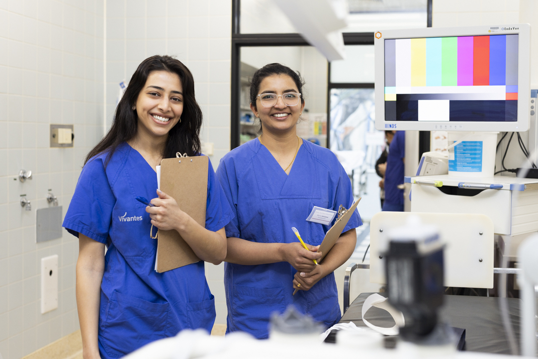 The two HPI master's students Eva Habib (left) and Sharmy Ann James smile friendly into the camera. They are in a room at Vivantes Hospital, wearing blue lab coats and holding clipboards in their hands. 