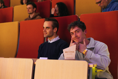 Niklas Kämmer zeigt seinen „Best Pitch“-Preis und zwei Daumen nach oben von den Sitzreihen im Hörsaal
