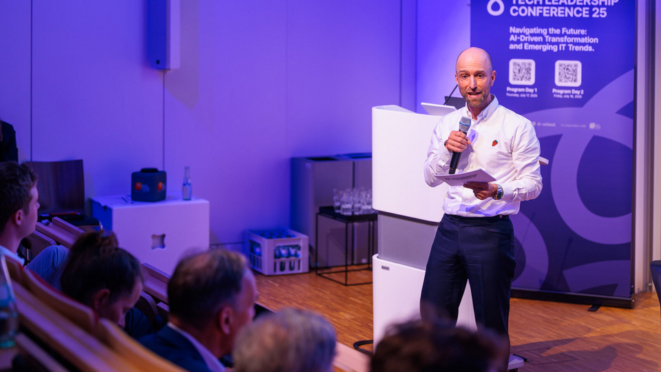 A man speaks into a microphone in front of an audience at the Tech Leadership Conference 2025. Behind him, a banner displays the conference title: “Navigating the Future: AI-Driven Transformation and Emerging IT Trends”