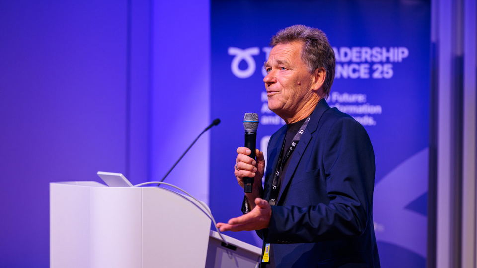 An older man speaks on stage at the Tech Leadership Conference 25. He holds a microphone in one hand and gestures with the other, standing at a podium with a blue conference banner in the background.