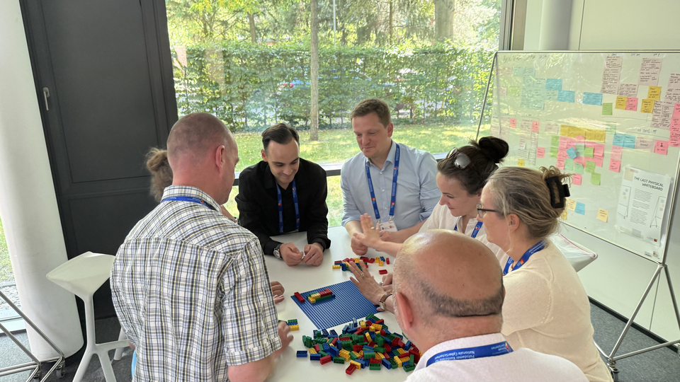 Workshop-Teilnehmende sitzen um einen Tisch mit bunten LEGO-Steinen und besprechen gemeinsam den Bau eines Prototyps; daneben steht ein Whiteboard mit Haftnotizen.