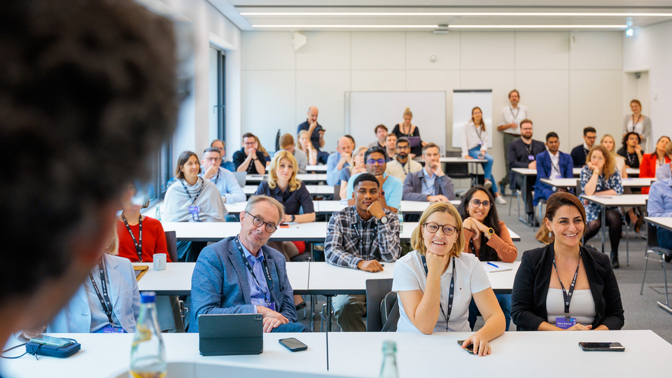 A speaker (visible only from behind) addresses a large and attentive audience in a modern workshop room during the Tech Leadership Conference 2025. The participants look engaged, some smiling or taking notes.