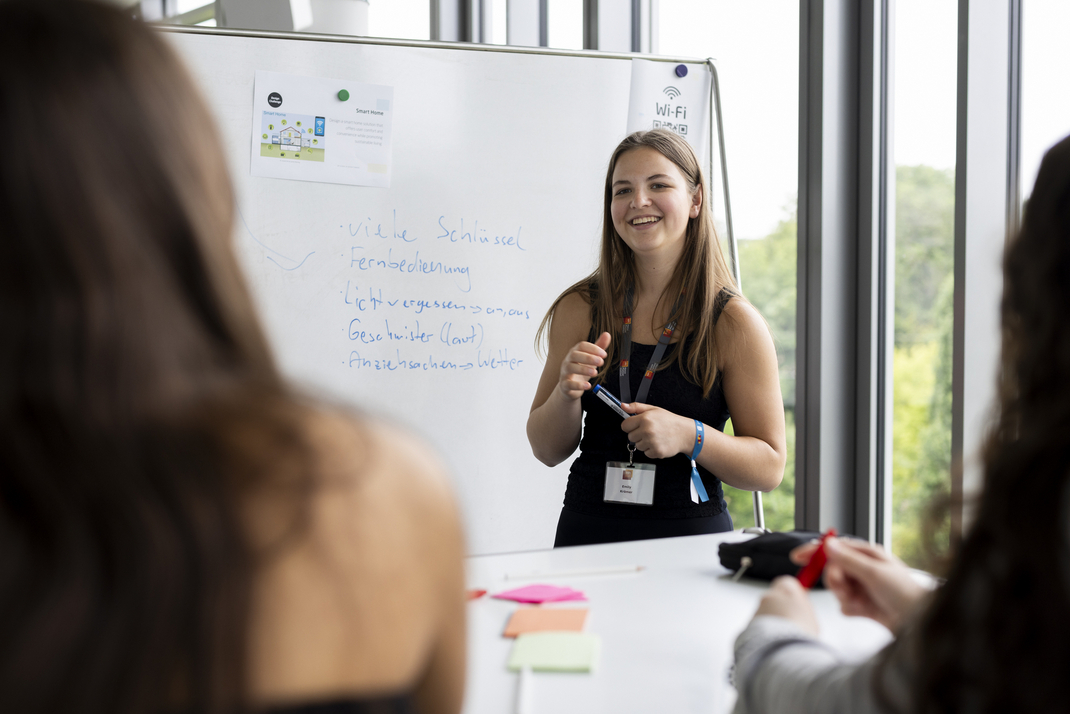 Teilnehmerinnen sitzen an einem Tisch mit einer Vielfalt an Schreibmaterialien und beraten sich untereinander. Eine Teilnehmerin schreibt Ergebnisse an ein Whiteboard 
