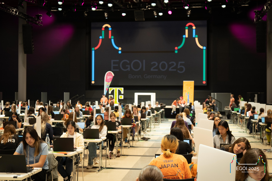 Die Teilnehmerinnen der EGOI absolvieren in einer Halle komplexe Prüfungen an Einzelplätzen. Im Hintergrund ist eine Leinwand mit dem Text "EGOI 2025 Bonn, Germany" zu sehen.