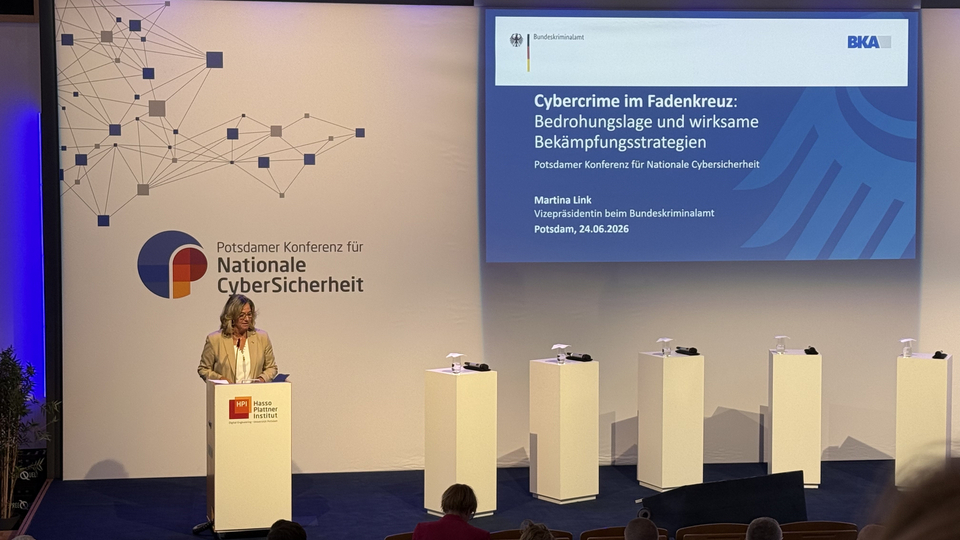 Eine Rednerin steht bei einer Cybersecurity-Konferenz am Rednerpult; auf der großen Leinwand ist eine Folie zu Cybercrime-Bedrohungen und Bekämpfungsstrategien zu sehen.