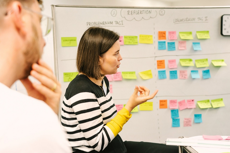 Several people are talking in front of a whiteboard with Post-it notes on it. 