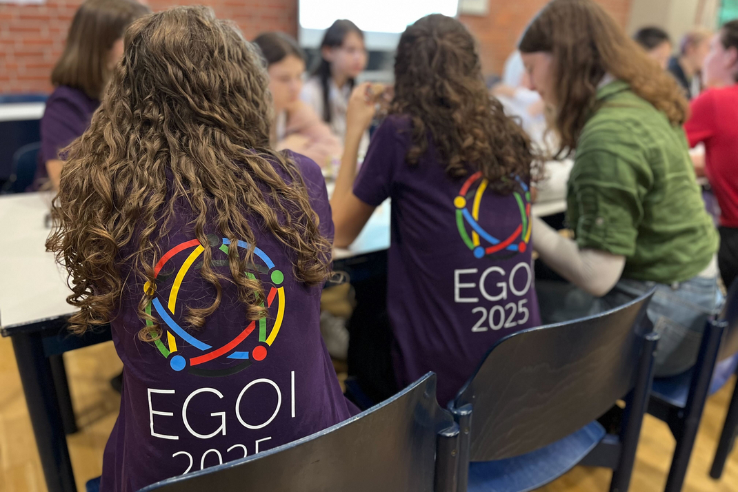 Mehrere Teilnehmerinnen der EGOI sitzen gemeinsam an einem Tisch. zwei von ihnen tragen ein EGOI-T-Shirt mit großem Logo auf dem Rücken.