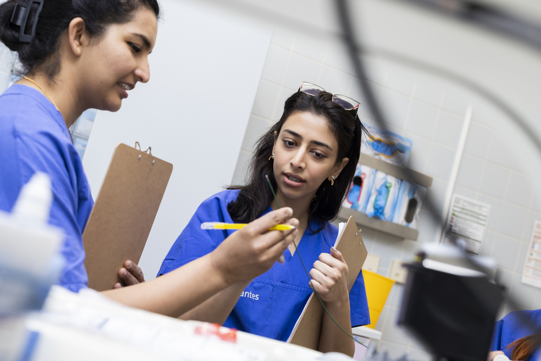 The two HPI master's students Eva Habib and Sharmy Ann James look with interest at a medical device. They are wearing blue lab coats and holding clipboards in their hands. 