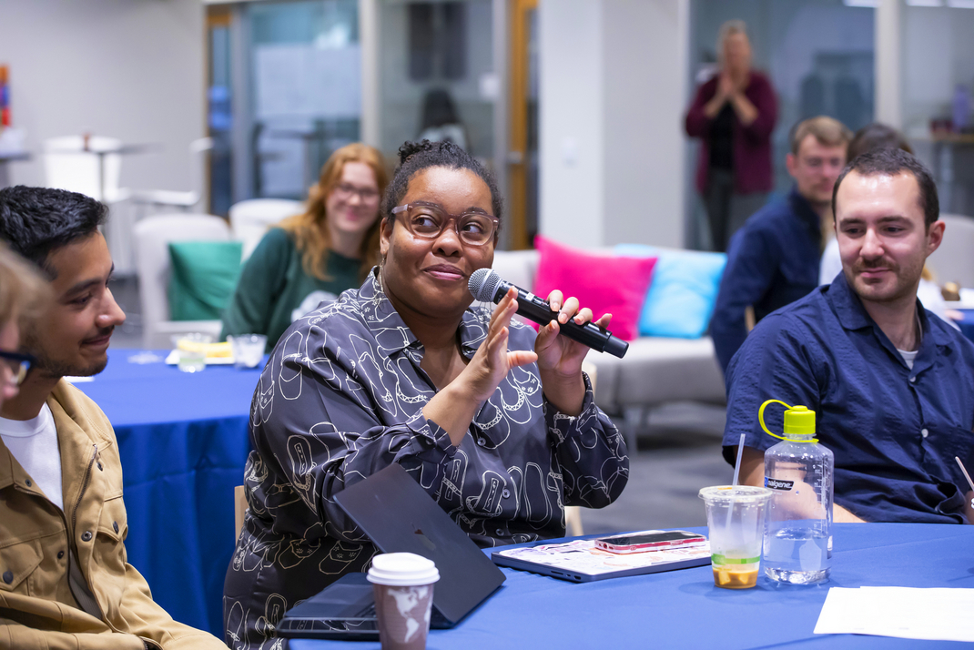 A workshop participant speaks into a microphone. She is seated at a round table with other workshop participants.