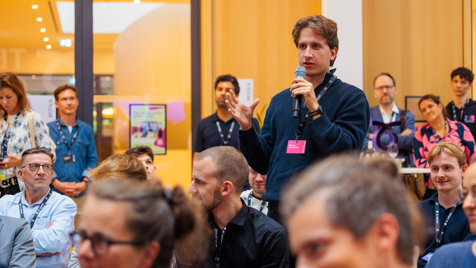 A young participant asks a question using a microphone during a discussion session at the Tech Leadership Conference 2025. He stands among the seated audience, who are listening attentively, while other attendees are visible in the background.