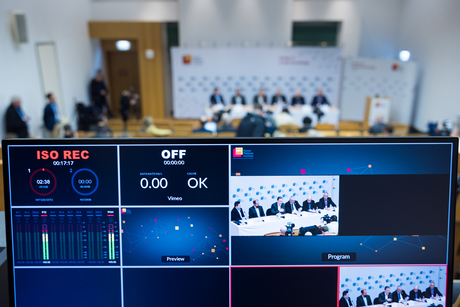 Im Vordergrund ist ein technischer Kontrollmonitor zu sehen, während im Hintergrund ist verschwommen zu erkennen ist, wie eine Konferenz vorbereitet wird.