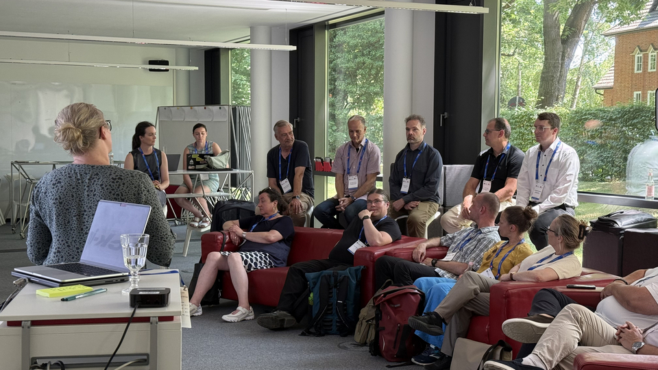 Teilnehmende sitzen in einem hellen Konferenzraum auf roten Sofas und Bänken und hören einer Moderatorin zu, die an einem Laptop präsentiert.