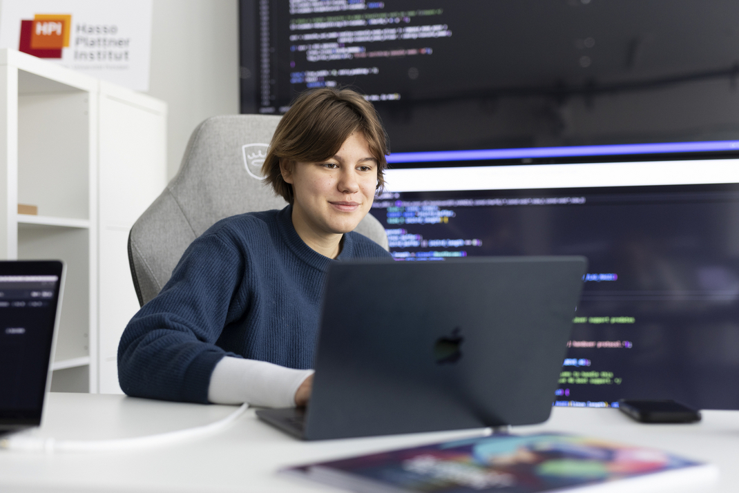 A person is working on a laptop in the office. Large displays with program code can be seen in the background