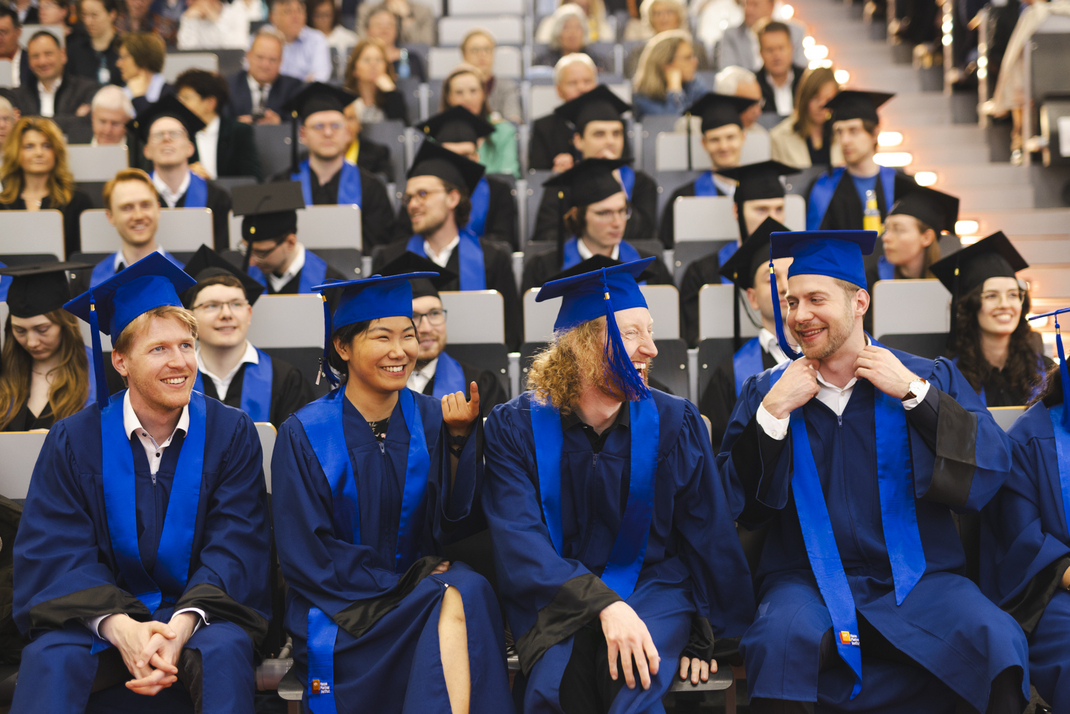 The photo shows graduates dressed in festive gowns, looking cheerful, in the audience during the ceremony