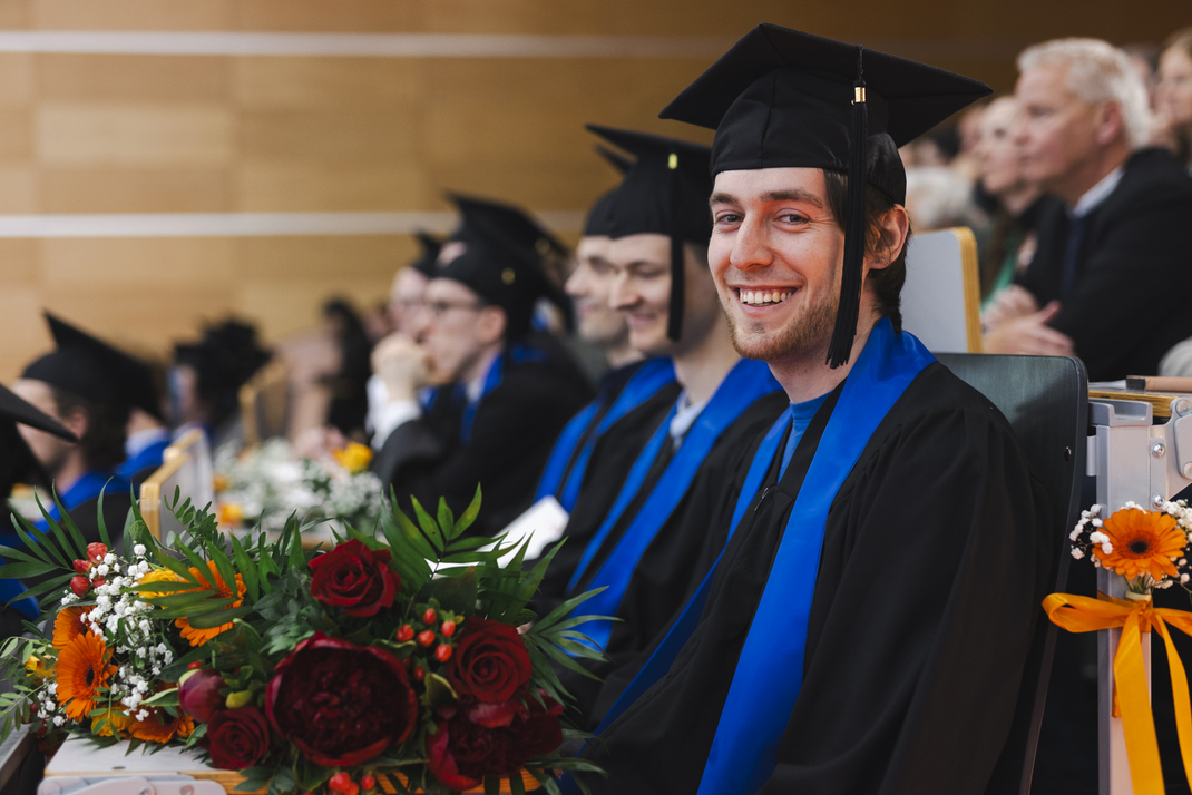 The photo shows proud graduates in the audience, holding their diplomas and bouquets of flowers