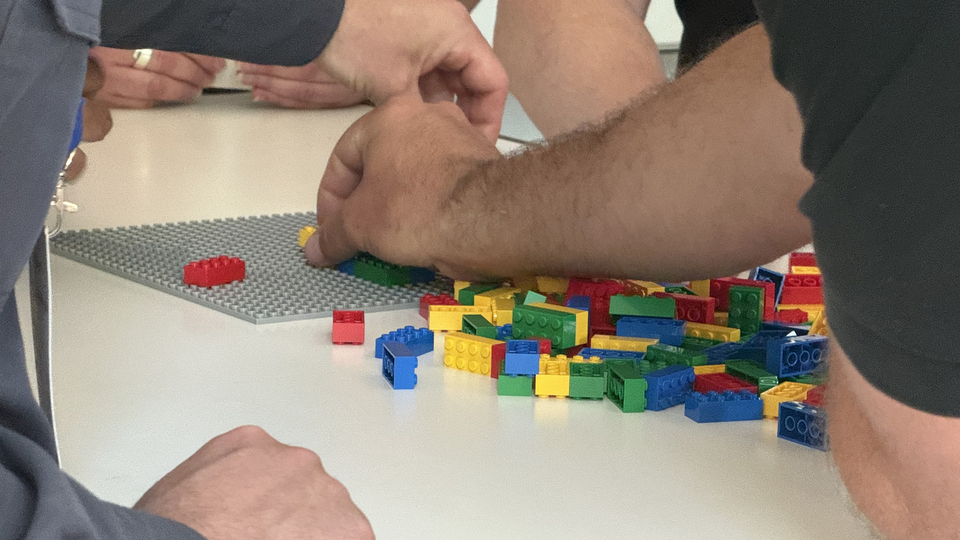 Nahaufnahme mehrerer Hände, die bei einer kollaborativen Prototyping-Übung bunte LEGO-Steine auf einer Grundplatte zusammensetzen.