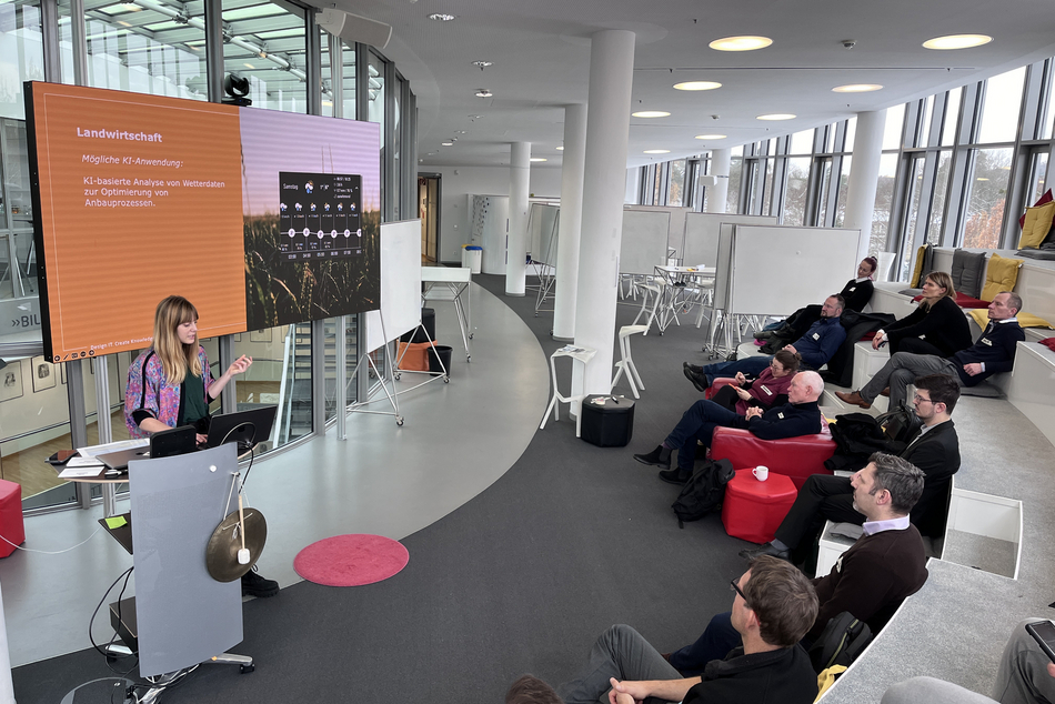 Woman behind a lectern with presentation, workshop participants seated opposite, listening.