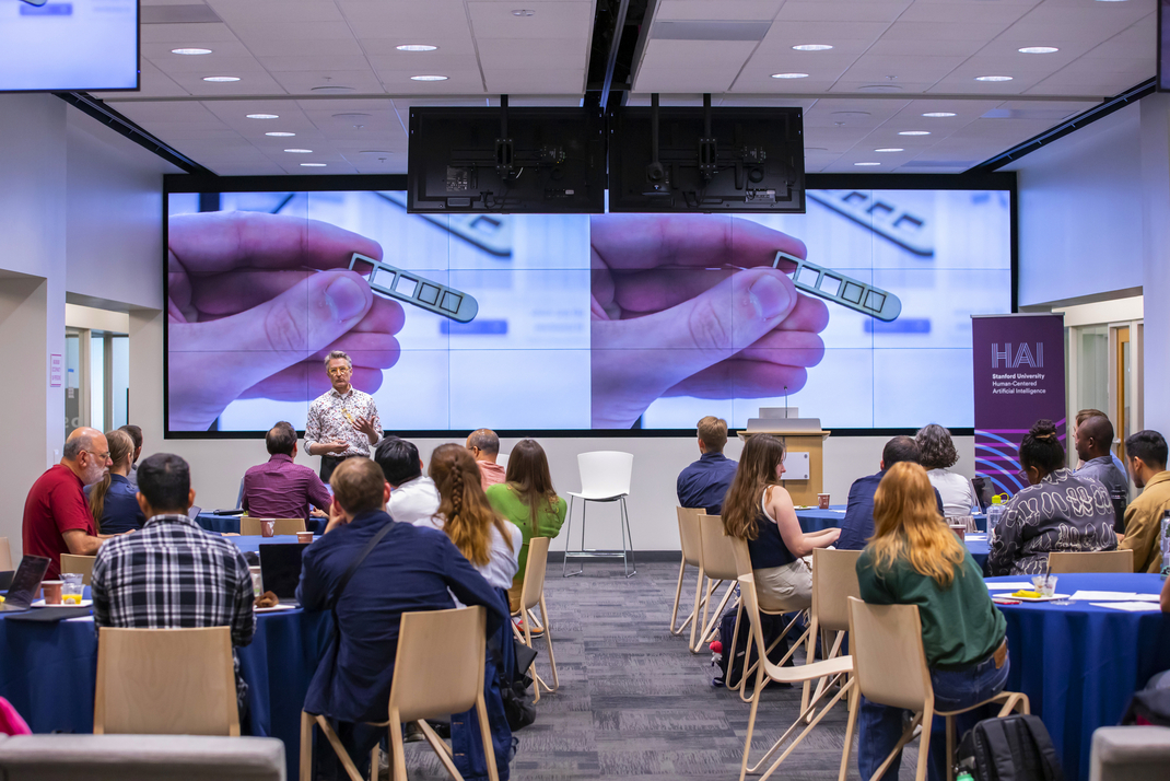 The workshop participants sit at round tables in a room. At the front, HPI professor Patrick Baudisch gives a lecture in front of a presentation.