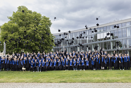 Graduates of the Hasso Plattner Institute toss their caps into the air on a lawn in front of the institute building