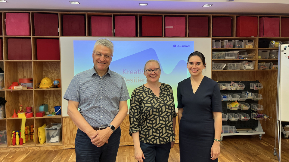 Drei Personen stehen lächelnd vor einer Präsentationsfolie mit dem Titel „Kreativität und Resilienz“ im Innovationsraum der HPI D-School. Im Hintergrund sind Regale mit buntem Kreativmaterial sichtbar.