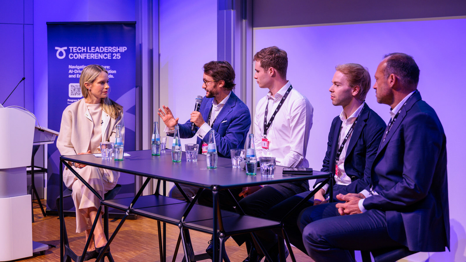 A panel discussion at the Tech Leadership Conference 25. A woman and four men sit at a table on stage, engaged in conversation. One man speaks into a microphone while the others listen attentively. Water bottles and glasses are placed on the table, and a conference banner is visible in the background.