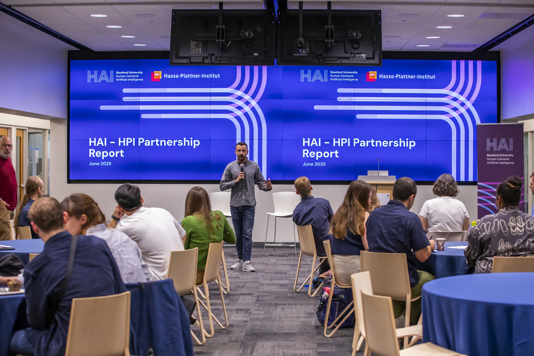 The workshop participants sit at round tables in a room. At the front, a lecturer presents a HAI-HPI Partnership Report, which can be seen in the background.