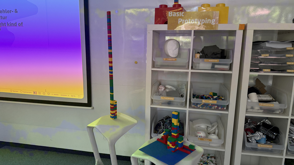 Ein Basic-Prototyping-Bereich mit Regalen voller Bastelmaterialien und LEGO-Steinen; davor stehen zwei hohe LEGO-Turmstrukturen auf kleinen Tischen.