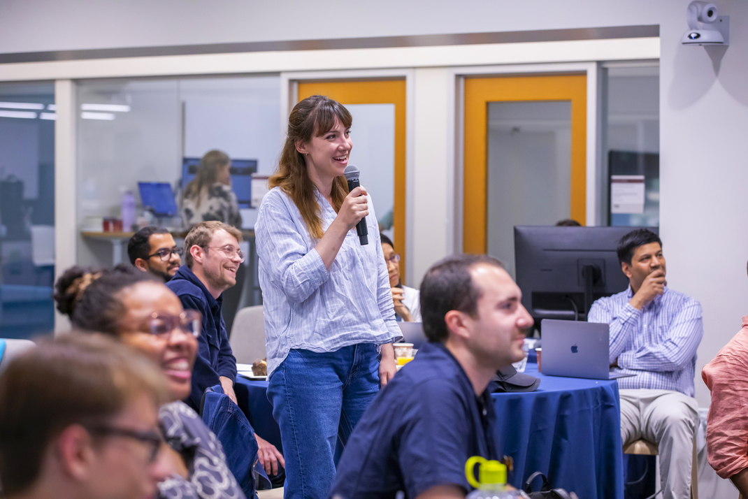 A workshop participant speaks into a microphone. Other workshop participants are seated around her.