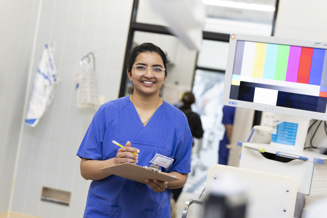HPI Master's student Sharmy Ann James in the Vivantes Clinic. She smiles friendly into the camera, wears a blue coat and holds a clipboard and a pen in her hands.