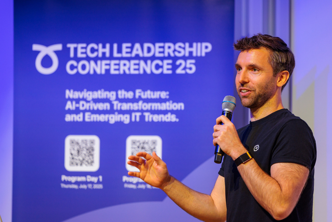 A speaker at the Tech Leadership Conference talks into a microphone. In the background, a banner advertising the conference can be seen.