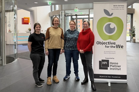 HPI student Lina Wilske, doctoral candidate Swantje Lange, Prof. Jiska Classen, and office assistant Mélanie Anoujard (from left to right) stand next to a banner for the Objective for the We conference in the lobby of the Hasso Plattner Institute.