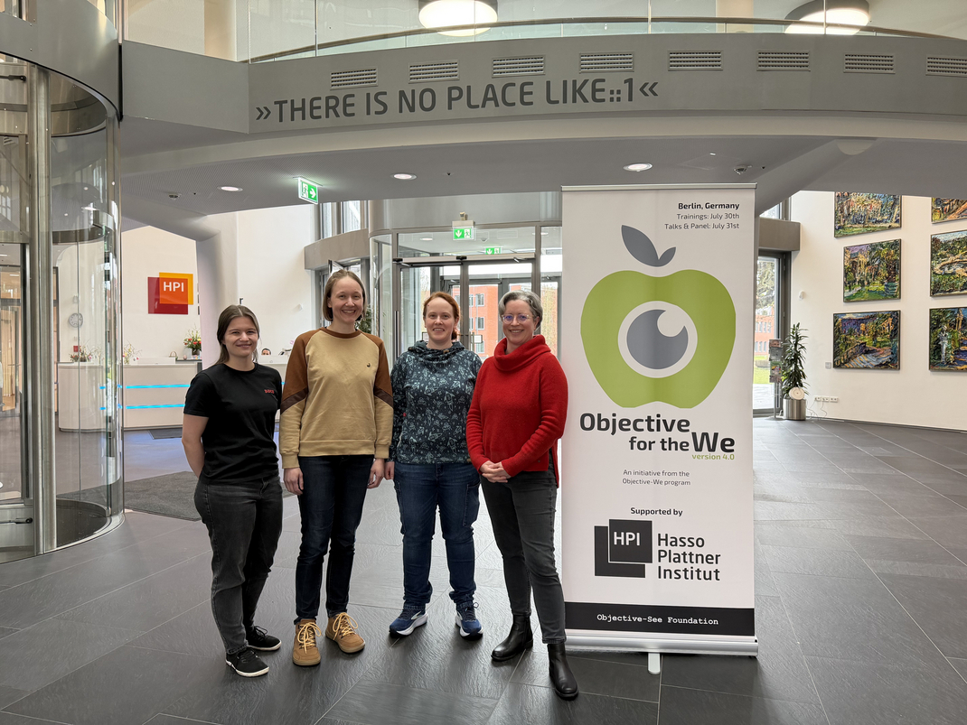 HPI student Lina Wilske, doctoral candidate Swantje Lange, Prof. Jiska Classen, and office assistant Mélanie Anoujard (from left to right) stand next to a banner for the Objective for the We conference in the lobby of the Hasso Plattner Institute.