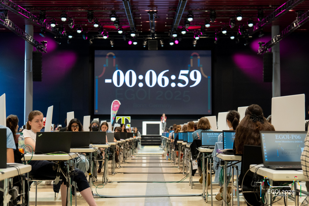 Die Teilnehmerinnen der EGOI absolvieren in einer Halle Prüfungen an Einzelplätzen. Auf der Leinwand im Hintergrund zeigt ein Timer "-00:06:59".