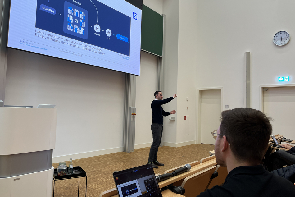 Speaker gestures in front of a presentation, foreground with audience members and laptops.