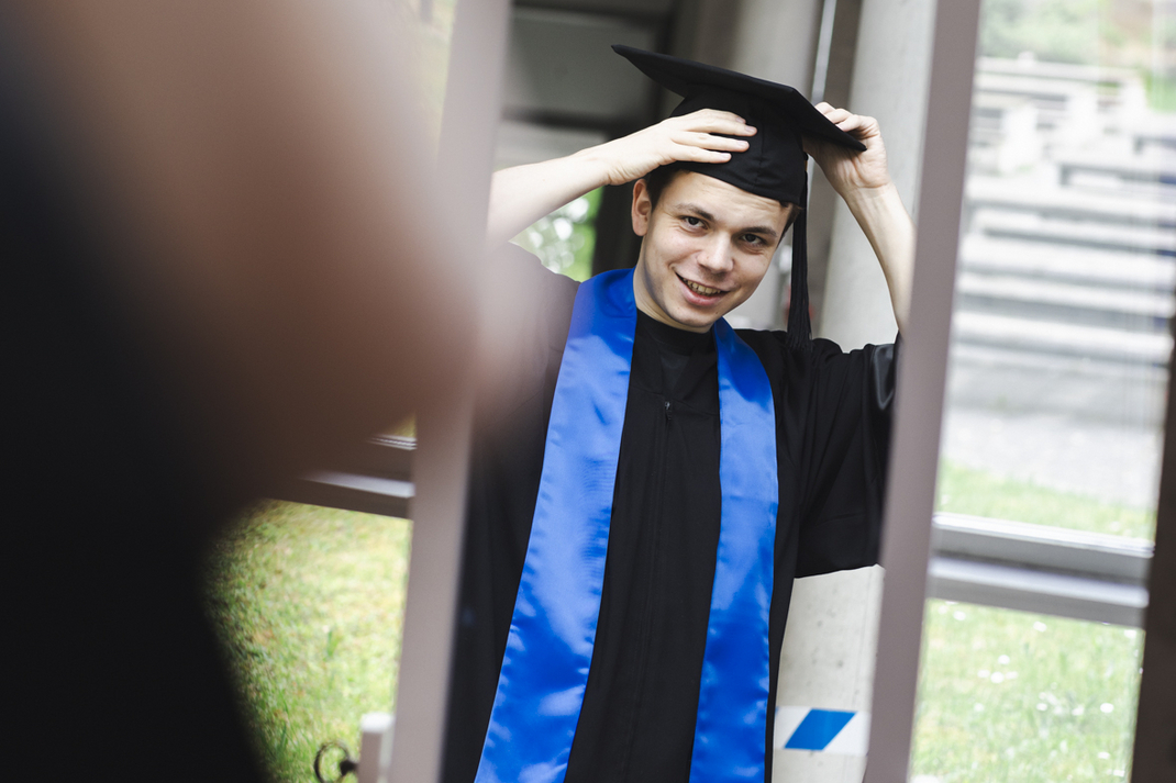 The photo shows a graduate looking at their graduation gown in the mirror