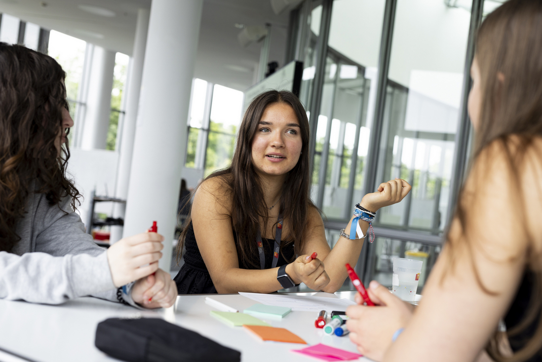Teilnehmerinnen sitzen an einem Tisch mit einer Vielfalt an Schreibmaterialien und beraten sich untereinander