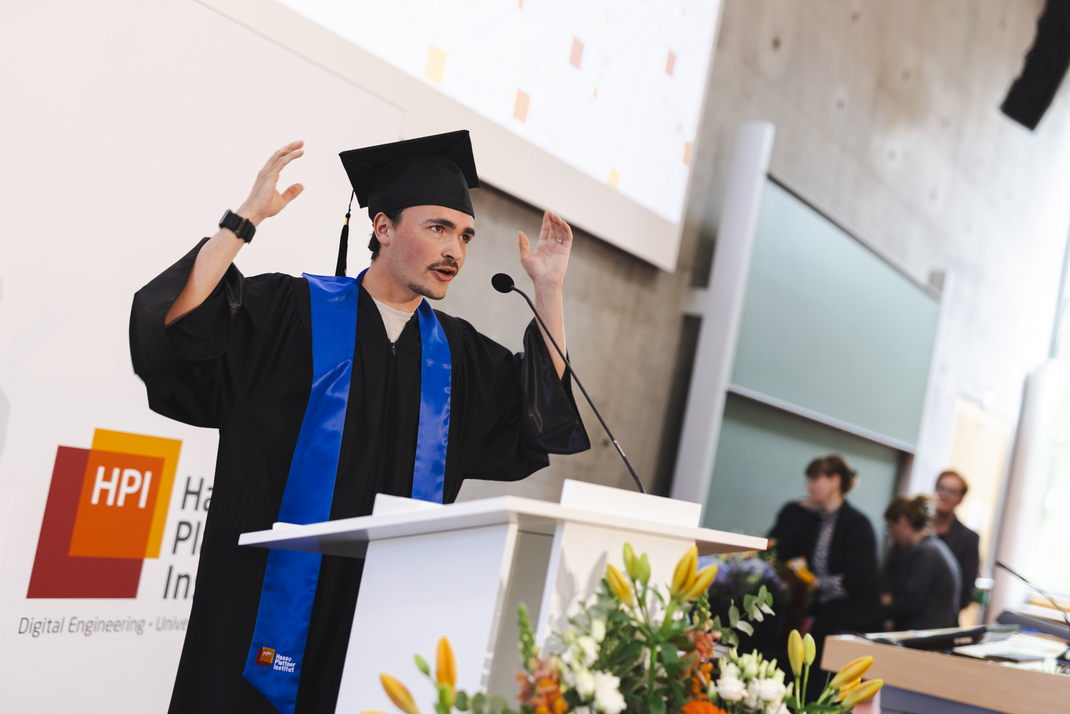 The photo shows an HPI graduate at the lectern. He throws his arms in the air as he speaks.
