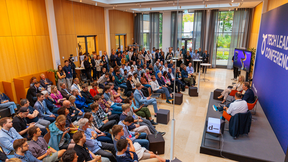 A large audience listens to a panel discussion at the Tech Leadership Conference, held in a bright room with a high ceiling and large windows. Two panelists sit on stage near a screen showing a fireplace image, while attendees sit on chairs and benches facing them.