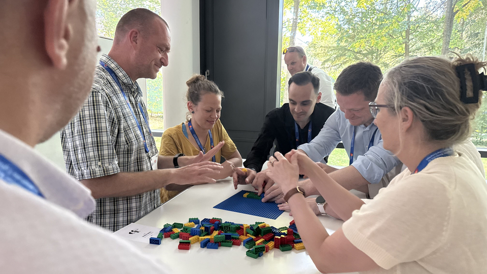 Eine Gruppe von Workshop-Teilnehmenden sitzt und steht um einen Tisch und baut gemeinsam eine kleine LEGO-Struktur auf einer blauen Grundplatte.