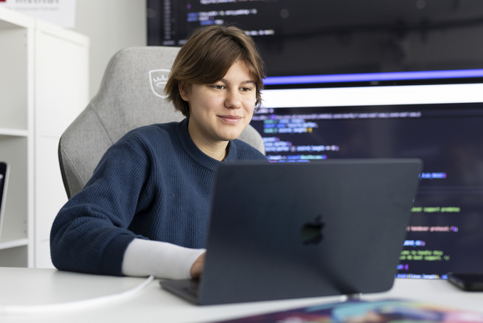 Eine person arbeitet im Büro an einem Laptop. Im Hintergrund sind große Displays mit Programmcode zu sehen.